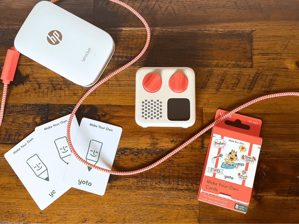 Image of a yoto mini player laying out along with three make your own cards, a yoto charger and a sprocket mini printer. 